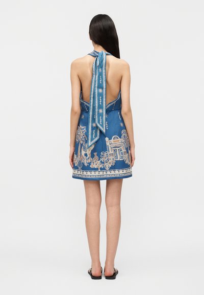 Γυναίκα με μακριά σκουρόχρωμα μαλλιά που φοράει γαλάζιο φόρεμα χωρίς πλάτη με μπεζ περίτεχνα σχέδια, στέκεται με τα χέρια στα πλάγια, έχοντας την πλάτη στραμμένη προς τα εμπρός.