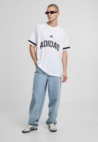 Bela majica s kratkimi rokavi in črnim logotipom Adidas ter črtastimi rokavi, v kombinaciji s svetlo modrimi širokimi kavbojkami in črnimi sončnimi očali.