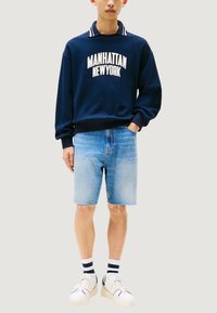 Felpa blu navy con scritta "MANHATTAN", abbinata a pantaloncini di denim azzurro chiaro, sneakers bianche e calzini a righe bianchi e neri.