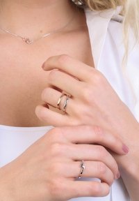 Two rings on hands: one smooth, polished silver band and one zigzag-shaped silver band. A delicate silver necklace with a heart accent is visible.