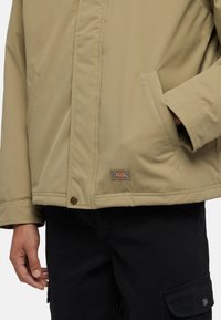 Beige insulated jacket with a zipper front, two side pockets, and a logo patch. Features stitched detailing and a relaxed fit.