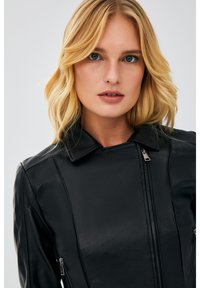 Black leather jacket featuring a zip front, stand collar, and side zip pockets. Smooth texture with vertical seam details.