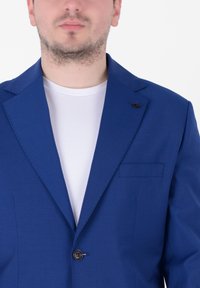 Blazer blu realizzato in tessuto liscio, con un solo bottone, revers a punta e taschino sul petto. Indossato sopra una maglietta bianca.