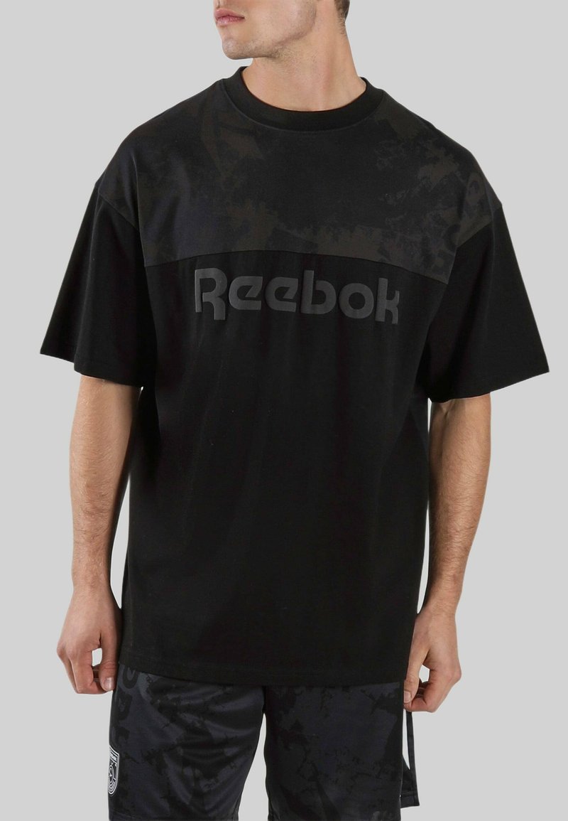 Hombre vistiendo una camiseta negra oversize de Reebok y pantalones cortos negros con estampado, de pie contra un fondo gris, mirando hacia un lado.