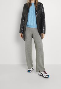 Černý lesklý kabát, světle modrý pletený svetr, šedé kalhoty s širokými nohavicemi a černobílé tenisky s modrými akcenty. Neutrální pozadí.