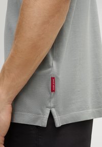 Lichtgrijze T-shirt met korte mouwen en een zijsplit, voorzien van een rood logo-label. De stof lijkt textuur te hebben, met zichtbare stiksels en een relaxte pasvorm.