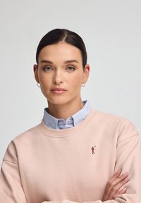 Ljusrosa sweatshirt med rund halsringning, prydd med en liten broderad logga i vinröd färg. Bars över en ljusblå skjorta med krage. Slät textur.