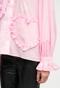 Ροζ μπλούζα με μακριά μανίκια, μανσέτες με βολάν και ένα μεγάλο, καρδιόσχημο μπαλώμα με βολάν στο μπροστινό μέρος, συνδυασμένη με μαύρο παντελόνι.