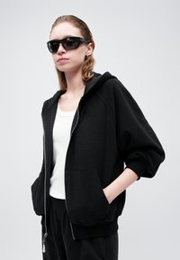 Svart zip-up hettegenser i teksturert stoff med avslappet passform, frontlommer og hette. Kombinert med en hvit tanktop og svarte joggebukser.