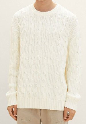 Mężczyzna ubrany w biały, gruby, pleciony sweter z długimi rękawami, zestawiony z beżowymi spodniami na jednolitym tle.