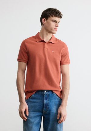 Mladý muž v hrdzavo sfarbenom polo tričku a modrých džínsach, stojaci a pozerajúci sa do strany na čistom bielom pozadí.