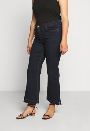Vrouw draagt donkerblauwe cropped wijde jeans, zwart mouwloos top en zwarte sandalen met lage hak en riempjes, staand op een grijze vloer.