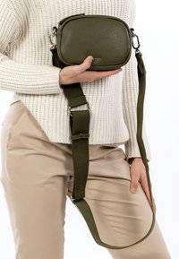 Alyvuogių žalsvos odos rankinė per petį su tekstūruota paviršiaus apdaila ir reguliuojamu audiniu dirželiu. Turi sidabro spalvos aparatūrą ir užtrauktuko uždarymą.