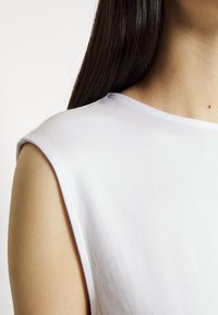 Femme aux longs cheveux foncés portant un haut blanc sans manches, montrant les détails des épaules et de l'encolure.
