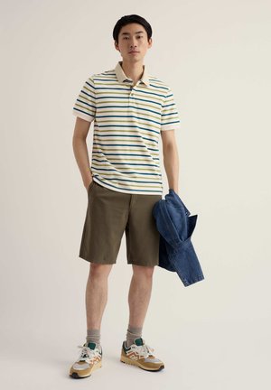 Junger Mann steht und hält eine blaue Jacke, trägt ein gestreiftes Poloshirt, olivgrüne Shorts, graue Socken und mehrfarbige Turnschuhe vor schlichter Hintergrund.