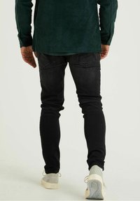 Jeans negros ajustados con un corte elegante, combinados con una camisa de pana verde oscuro texturizada. Zapatillas blancas con suelas claras completan el look.