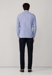 Uomo in piedi di fronte a un muro, indossa una camicia a maniche lunghe azzurra chiara, jeans blu scuro e scarpe nere con i risvolti alzati.