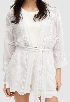 Femme portant une robe kimono en dentelle blanche transparente par-dessus un top blanc et un short à ourlet festonné, nouée à la taille avec une ceinture en tissu.