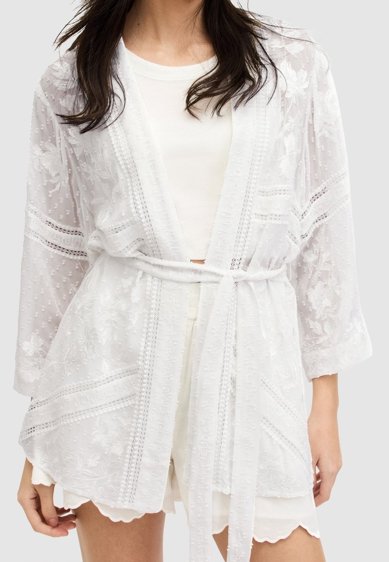 Femme portant une robe kimono en dentelle blanche transparente par-dessus un top blanc et un short à ourlet festonné, nouée à la taille avec une ceinture en tissu.
