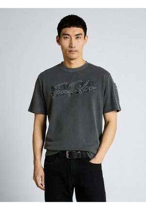 Man draagt een donkergrijs T-shirt met korte mouwen met de tekst "Tom Tailor", een zwarte broek en een zwarte riem, staand tegen een effen lichte achtergrond.