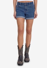 Denim šorti ar salocītām malām, klasiskā piecu kabatu dizainā, zilā krāsā, apvienoti ar pelēkām un melnām rietumu stila zābakiem ar sarežģītu dūrušanos.