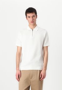 Hvit kortermet polo skjorte med teksturert overflate, glidelås i halsen og kontrastkrage. Kombinert med beige bukser.
