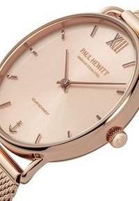Roségoldene Armbanduhr mit einem hellpinkfarbenen Zifferblatt, schlanken Stundenmarkierungen und römischer Ziffer XII. Sie verfügt über ein gewebtes Metallarmband und ein Ankerlogo.