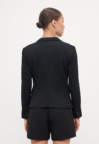 Svart blazer med slagkrage, skräddarsydd passform, texturerad tyg, lång ärm med knappdetalj, parat med svarta shorts. Bakvy visas.