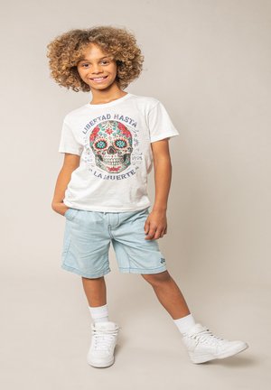 Enfant souriant aux cheveux bouclés portant un t-shirt blanc avec un imprimé crâne, un short bleu clair, des chaussettes blanches et des baskets montantes blanches, en position debout.