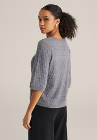 Grijze gebreide trui met korte mouwen, voorzien van een gestructureerd ruitpatroon. Het materiaal ziet er zacht en licht pluizig uit.