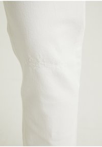 Pantalones de mezclilla blancos con corte recto, que presentan un parche desgastado en la rodilla izquierda y una tela suave y texturizada.