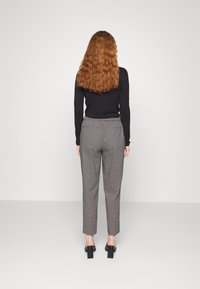 Woman with long curly red hair stands facing a white wall, wearing a black long-sleeve top, gray checkered pants, and black block heel shoes.