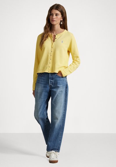 Yellow button-up cardigan made of soft fabric, paired with light blue loose-fit jeans. Features a small logo and round neckline.