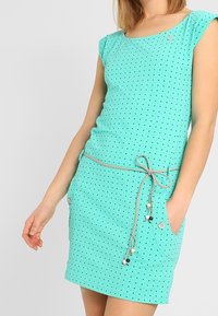 Ljust turkos klänning med korta ärmar, svarta prickar, resårmidja med snörning och sidofickor med knappaccent.