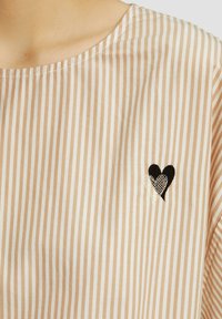 Top in tessuto leggero a righe in beige e crema con scollatura ampia. Presenta un piccolo cuore ricamato in bianco e nero sul petto.