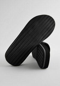 Schwarze Slipper mit einer strukturierten, gezackten Sohle. Das Obermaterial besteht aus weichem Stoff mit dezenten schwarzen und grauen Streifen; niedriges Profil.