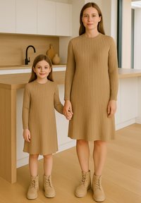 Zwei Figuren in gerippten, beigen Strickkleidern stehen nebeneinander und halten sich die Hände. Beide tragen beige Schnürstiefel. Eine moderne Küche ist im Hintergrund zu sehen.