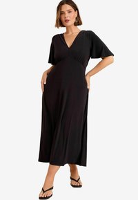 Robe noire fluide en tissu doux avec un col en V et de courtes manches volantées. Comprend des poches latérales et une coupe décontractée atteignant le milieu du mollet.