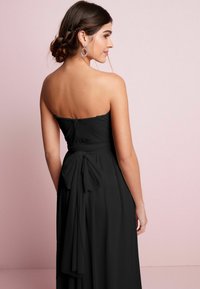 Musta olkaimeton mekko, jossa on tiukka bodice, virtaava hame ja suuri solki vyötäröllä. Takaa näkyy sileä, teksturoidun näköinen kangas.