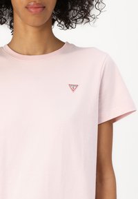 Tricou din bumbac roz deschis cu guler rotund și mâneci scurte, având un mic logo triunghiular "GUESS" în roșu și alb pe pieptul stâng.