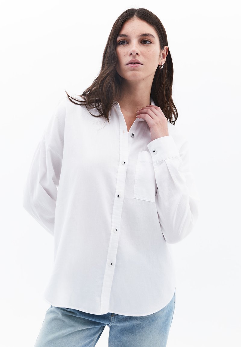 Camicia bianca a maniche lunghe con bottoni, dotata di tasca, bottoni neri e una vestibilità rilassata. Tessuto morbido, orlo curvo e texture delicata.