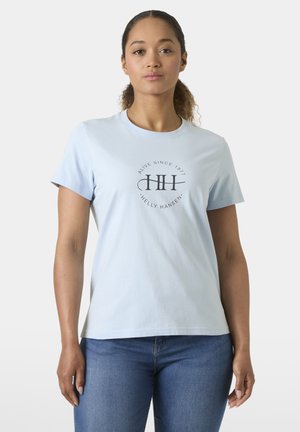 Kobieta o kręconych włosach, ubrana w jasnoniebieską koszulkę Helly Hansen i niebieskie dżinsy, stojąca na jednolitym jasnym tle.