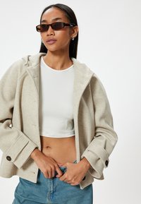Beige ullblandad jacka med huva, stora knappdetaljer, vida ärmar, bärs över en vit kort topp och blå högmidjade denimjeans.