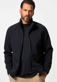 Chaqueta de cremallera azul marino con cuello alto, hecha de una tela suave. Presenta una cremallera frontal completa y dos bolsillos laterales, superpuesta sobre una camiseta negra.