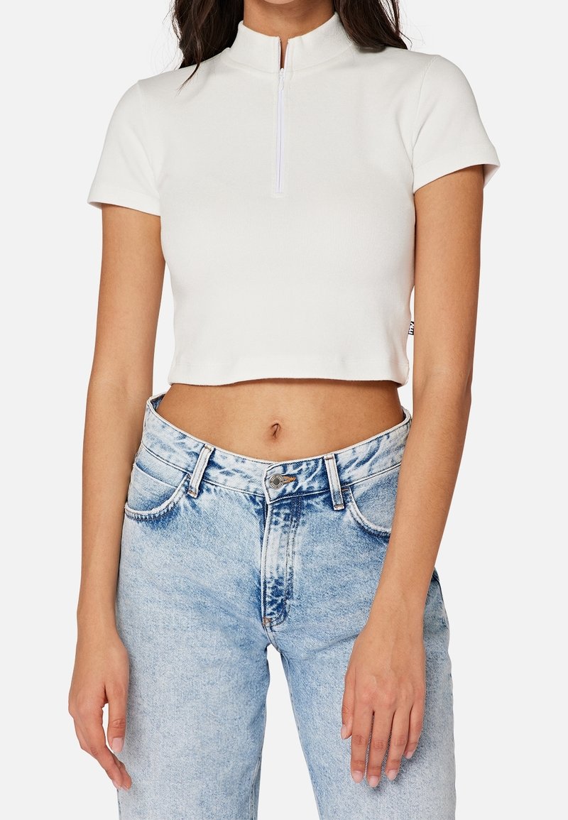 Haut blanc court avec un col montant et une fermeture éclair, fabriqué à partir d'un tissu lisse ; associé à un jean en denim bleu clair taille haute.