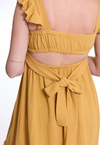 Senfgelbes Kleid mit Rüschenträgern, gerafftem Oberteil und offenem Rücken. Verfügt über einen gebundenen Taillenbund und einen strukturierten Stoff mit weichem Finish.