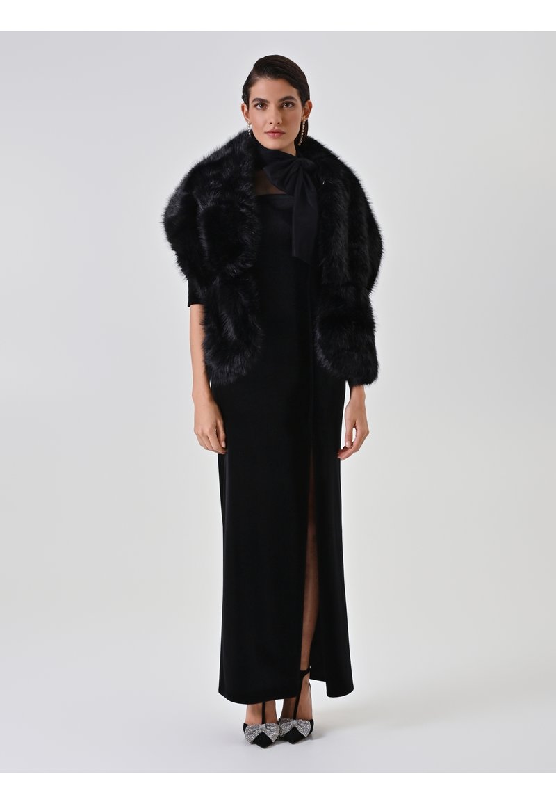 Robe de soirée noire avec une fente haute sur la cuisse, accompagnée d'un châle en fausse fourrure noire. Des talons scintillants noirs et argent complètent la tenue.