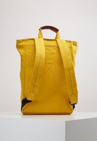 Zaino in tessuto giallo con design strutturato, dotato di spallacci doppi, una maniglia marrone e cuciture accennate sui lati.