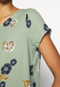 Květinová blůza z jemného zeleného materiálu s krátkým rukávem. Obsahuje květinové vzory v modré, béžové a oranžové barvě. Měkká, splývavá textura.