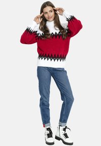 Kobieta w czerwonym, białym i czarnym swetrze w wzory, niebieskich dżinsach i białych wiązanych botkach, stojąca z rękami blisko twarzy.